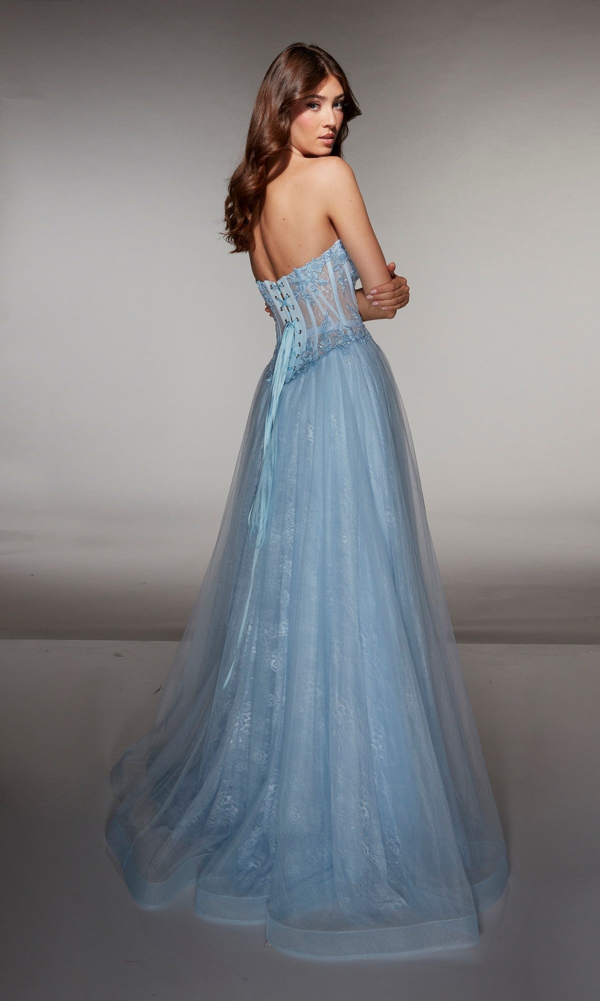 A woman stands with her back turned, wearing the Alyce 61984 long prom dress— a strapless light blue tulle gown with a corset bodice, lace-up back, and sheer details for an ethereal look.